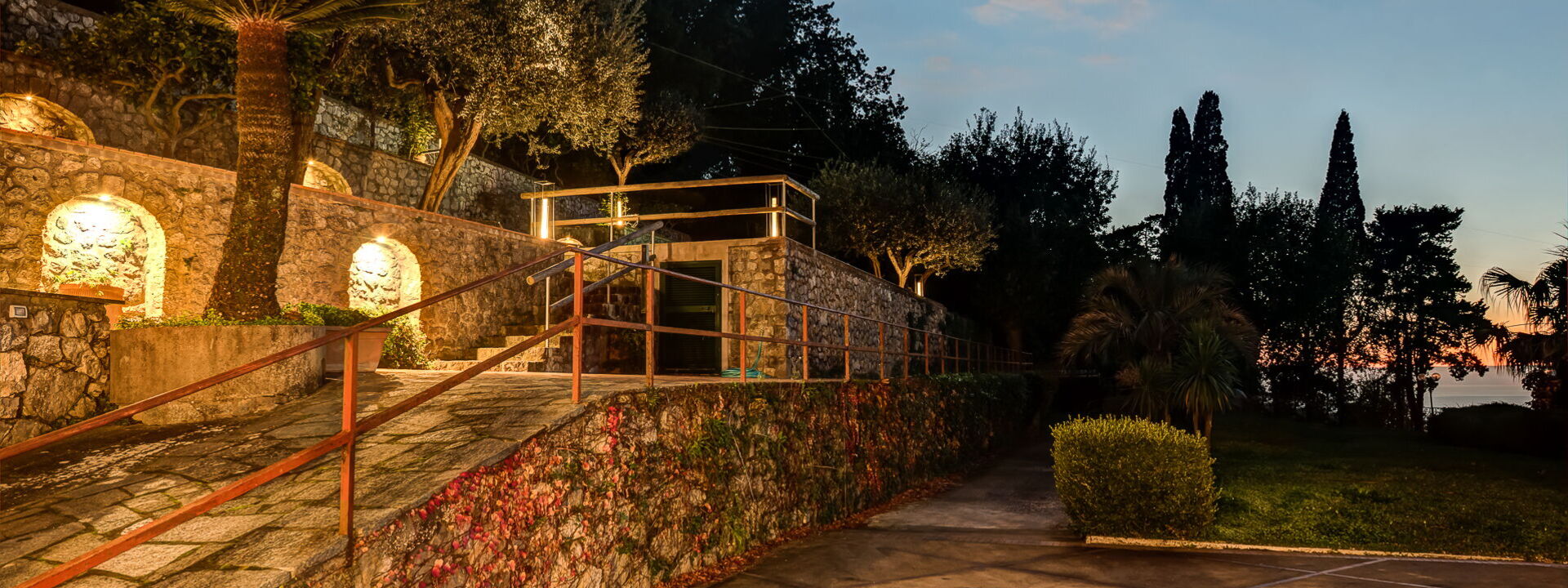 Il giardino di Villa Demetra al crepuscolo, con muri in pietra e alberi illuminati